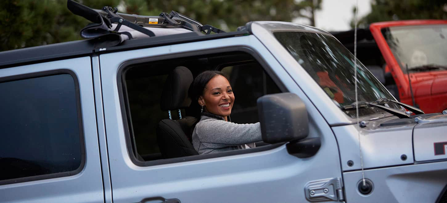 A woman smiling while driving a grey Jeep Wrangler with the windows down