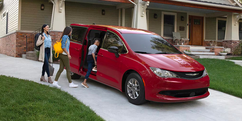 Children entering the rear passenger side of a red 2025 Chrysler Voyager parked in a residential driveway while a woman watches them enter the vehicle