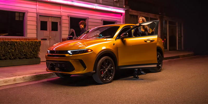 A vibrant yellow Dodge Hornet parked on a neon-lit city street with two people nearby.