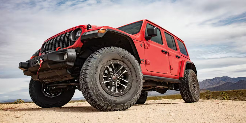 A red Jeep Wrangler with off-road tires, parked on a rugged desert terrain.