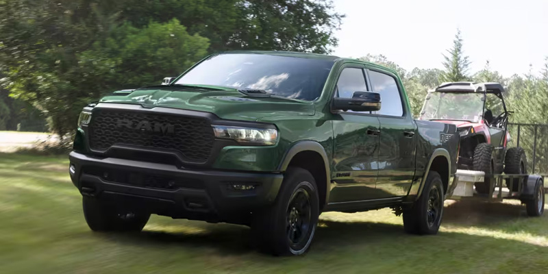 A 2026 RAM 1500 in green, towing an off-road vehicle on a trailer through a grassy area.