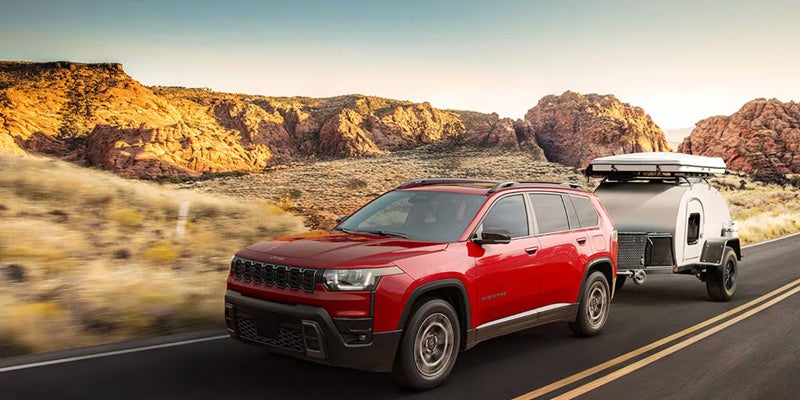 Red 2026 Jeep Cherokee towing a trailer on a desert road with rocky mountains in the background.
