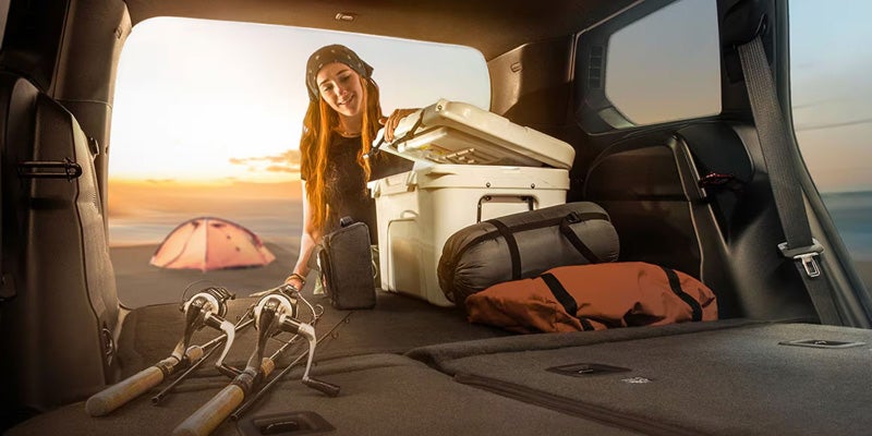 Woman loading camping gear into the rear cargo area of a 2026 Jeep Cherokee at sunset.