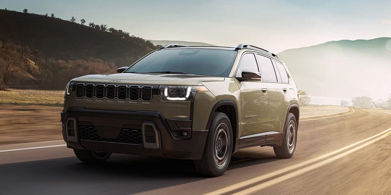 2026 Jeep Cherokee driving on a scenic road with mountains and water in the background.